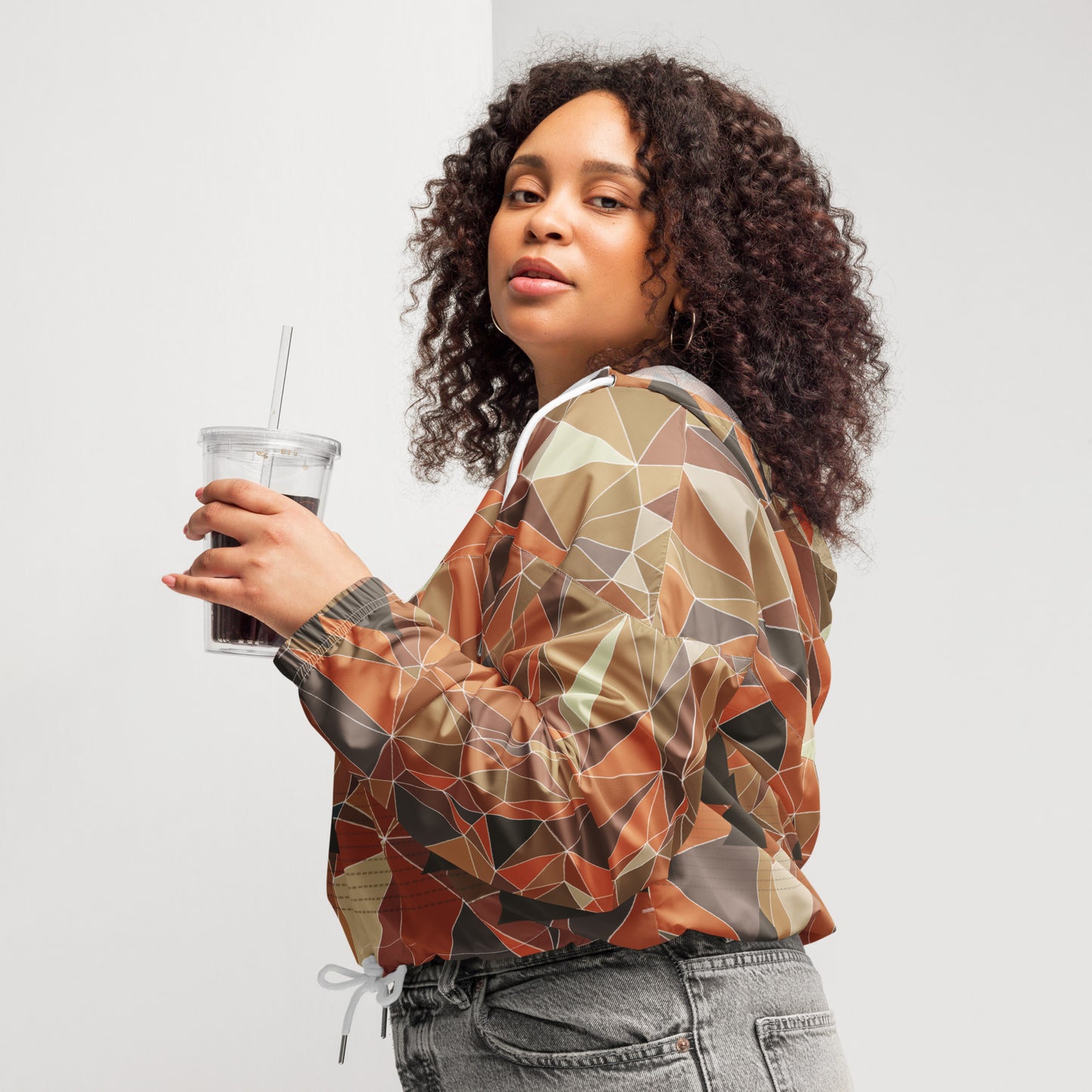 Woman wearing a cropped multicolor geometric patterned windbreaker jacket holding a drink against a plain background
